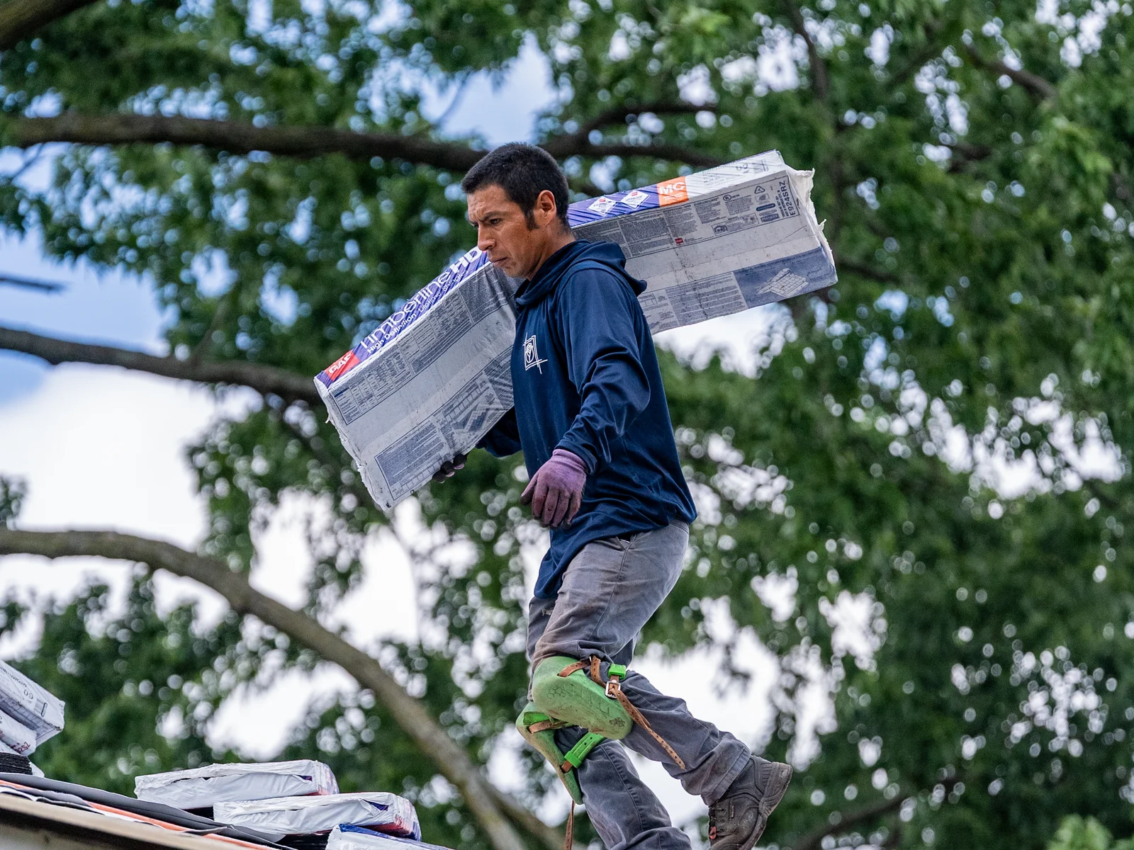 Rosales Roofing crew member carrying a shingle bundle along the roof edge past staged underlayment in Orland Hills, Illinois